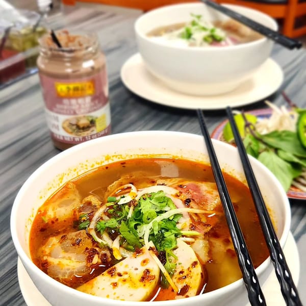 Two bowls of Vietnamese pho, one spicy, with chopsticks and a jar of chili sauce.