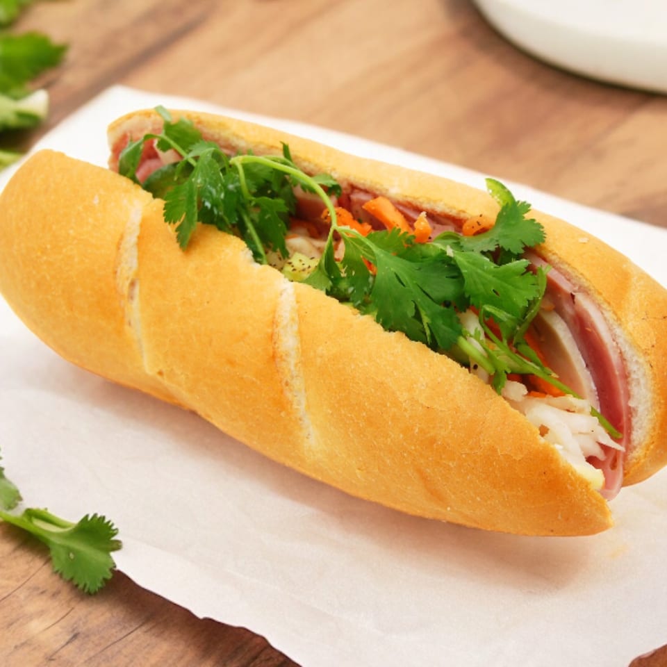 A Vietnamese banh mi sandwich filled with meats, pickled vegetables, and fresh cilantro on parchment paper.