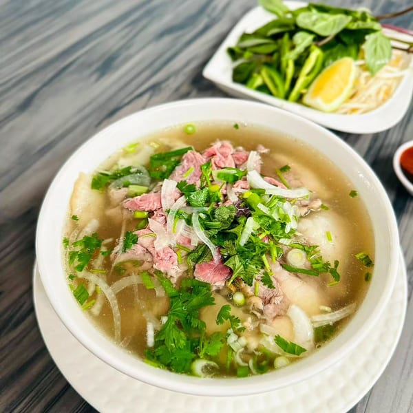 A bowl of Pho Huyen & Banh Mi's Vietnamese pho with beef, herbs, and onions, with a side of fresh garnishes.