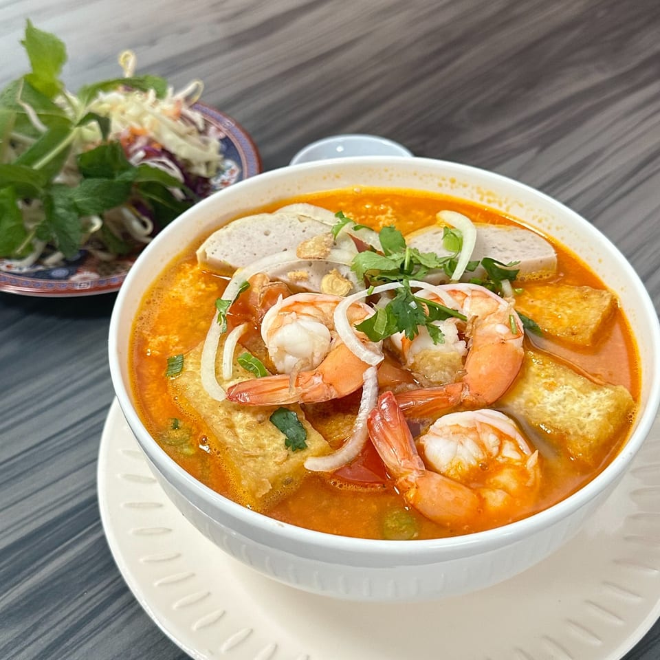 A vibrant bowl of Vietnamese spicy shrimp and tofu noodle soup with a side of fresh herbs.