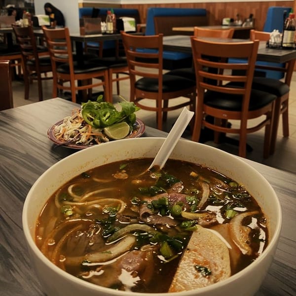 A large bowl of Vietnamese pho with a side of fresh herbs and lime in a restaurant setting.