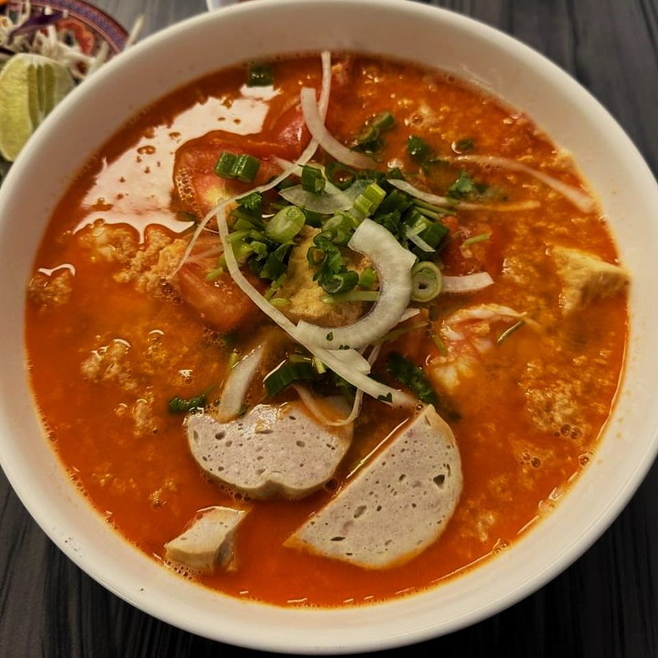 Close-up of a vibrant red Vietnamese soup with sliced sausage, tomatoes, and green onions.
